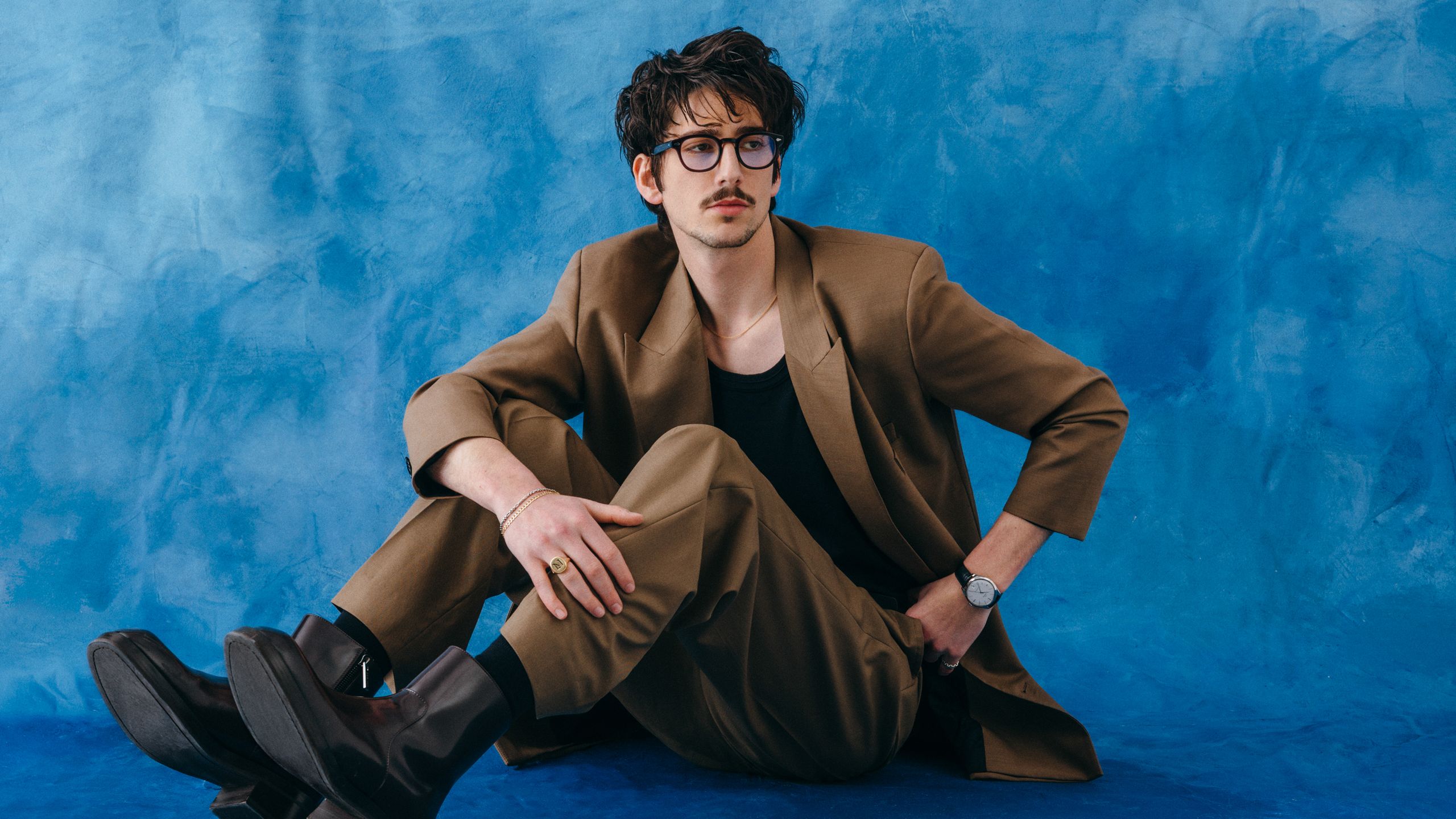 Milo wears glasses an unbuttoned tan suit black tank top and on watch on his left wrist while sitting on the blue...
