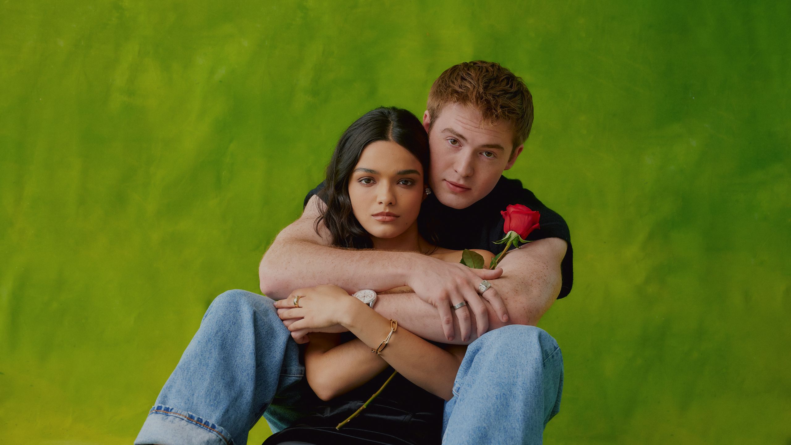 Kit Connor and Rachel Zegler against green painted backdrop. Both are sitting and Kit holds a red rose while his arms...